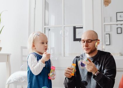 A joyful moment between a father and daughter blowing bubbles indoors, highlighting family fun.