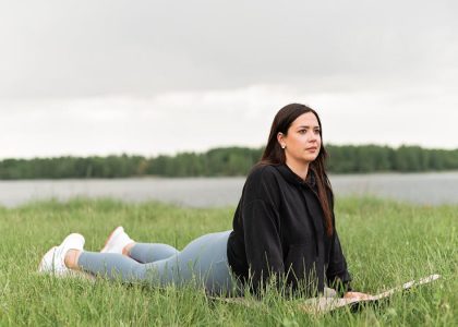 Woman lying on a yoga mat in a grassy field by a lake, promoting relaxation and wellness.